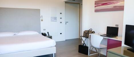 A modern hotel room with a bed, a desk with a laptop, a chair, a TV, and a painting on the wall.