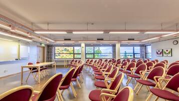 A conference room with rows of red chairs, a whiteboard, and a large window with a view of trees.