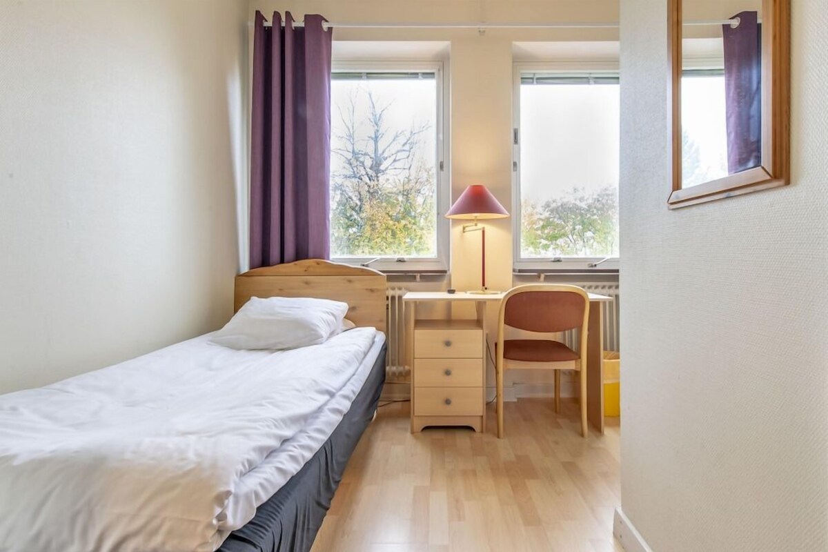 Een slaapkamer met een enkel bed, een bureau, een stoel en een raam met gordijnen.