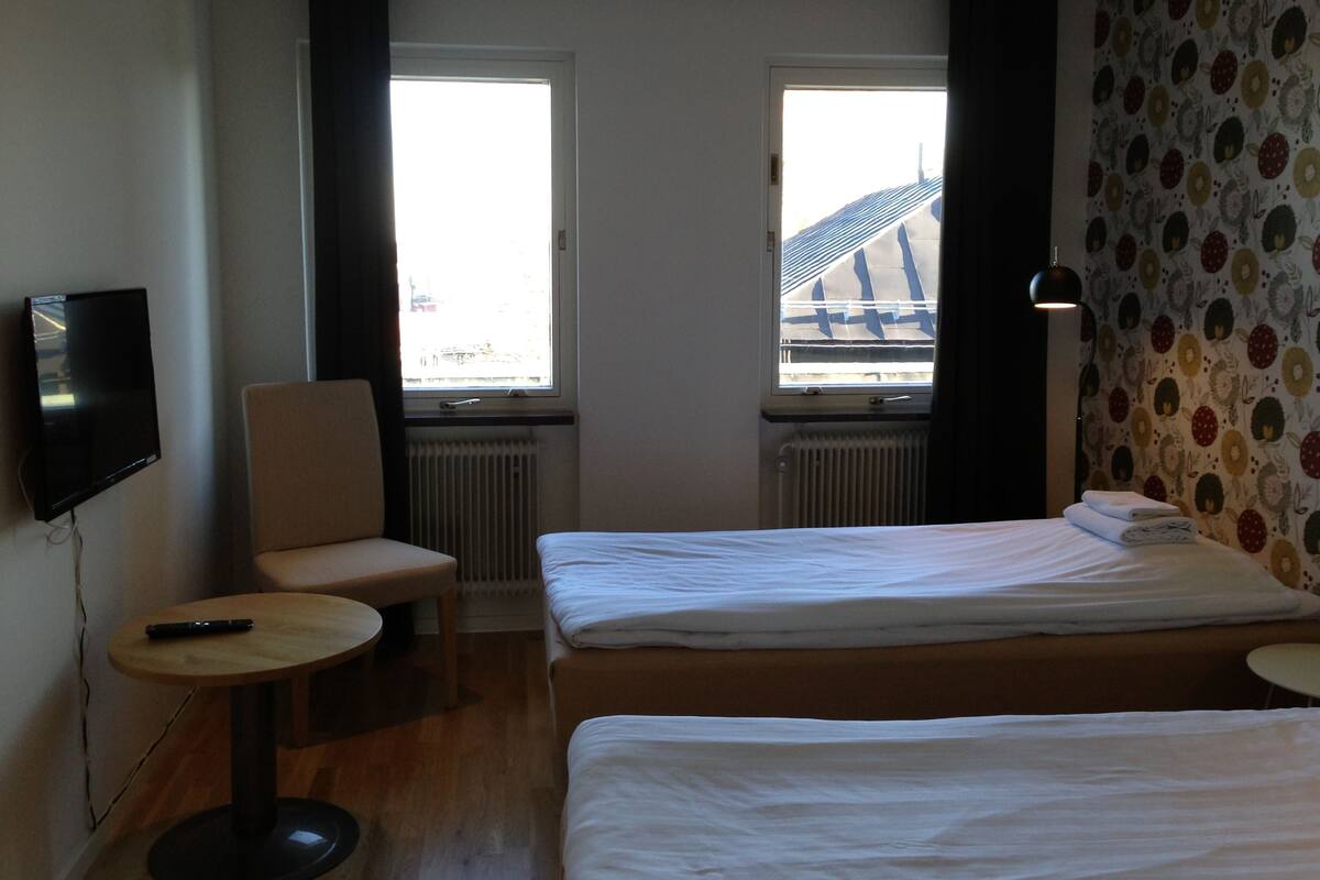 Hotellihuone, jossa on kaksi sänkyä, pieni pöytä, tuoli, televisio ja ikkuna, josta on näkymä rakennukseen.