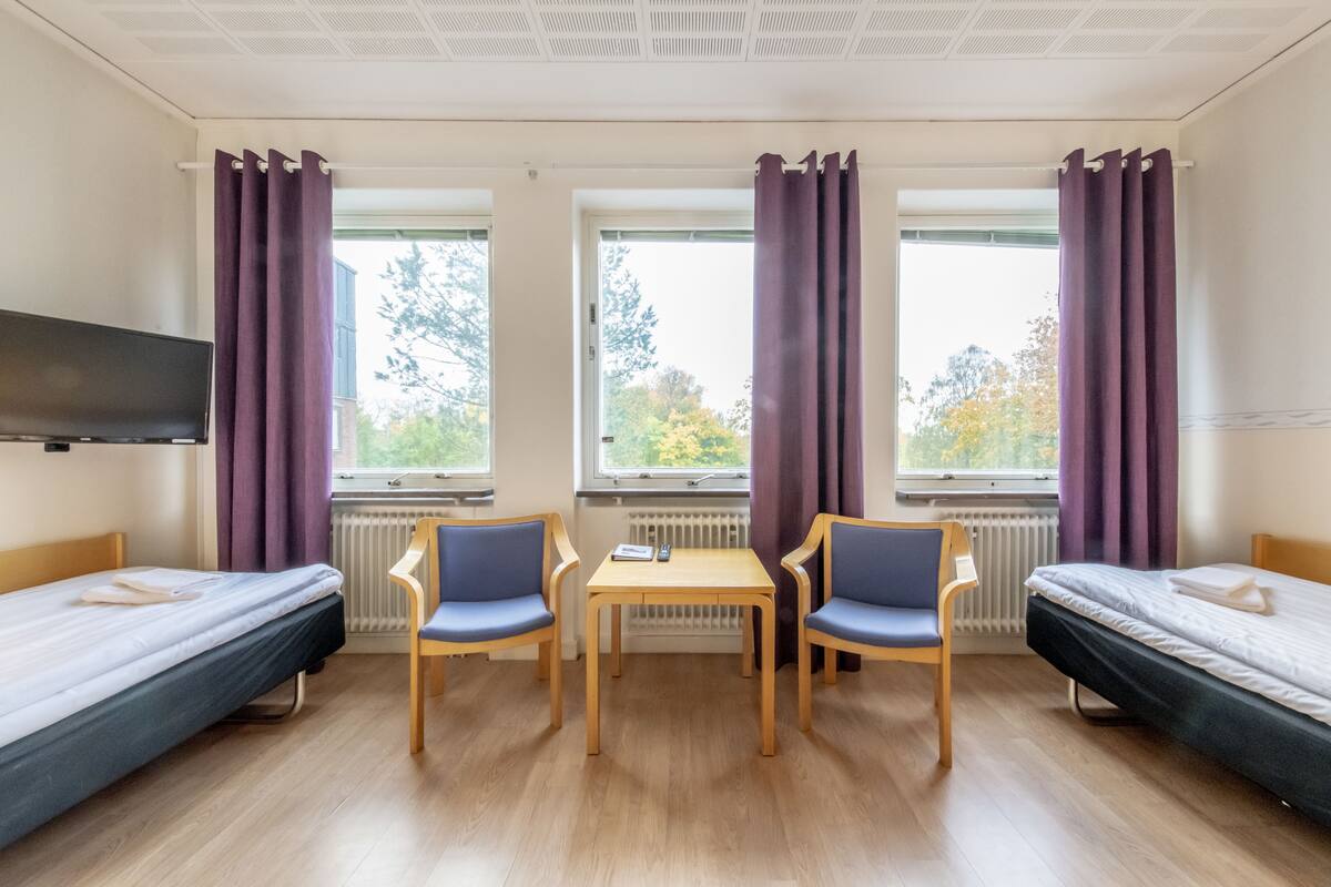 Huoneessa on kaksi erillistä sänkyä, pieni pöytä, kaksi tuolia, televisio ja suuret ikkunat, joissa on violetit verhot.