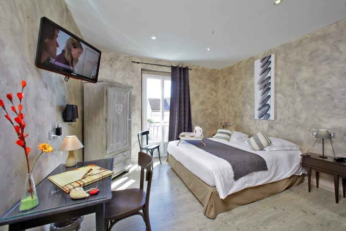 A hotel room with a bed, a desk with a chair, a TV mounted on the wall, and a window with curtains.