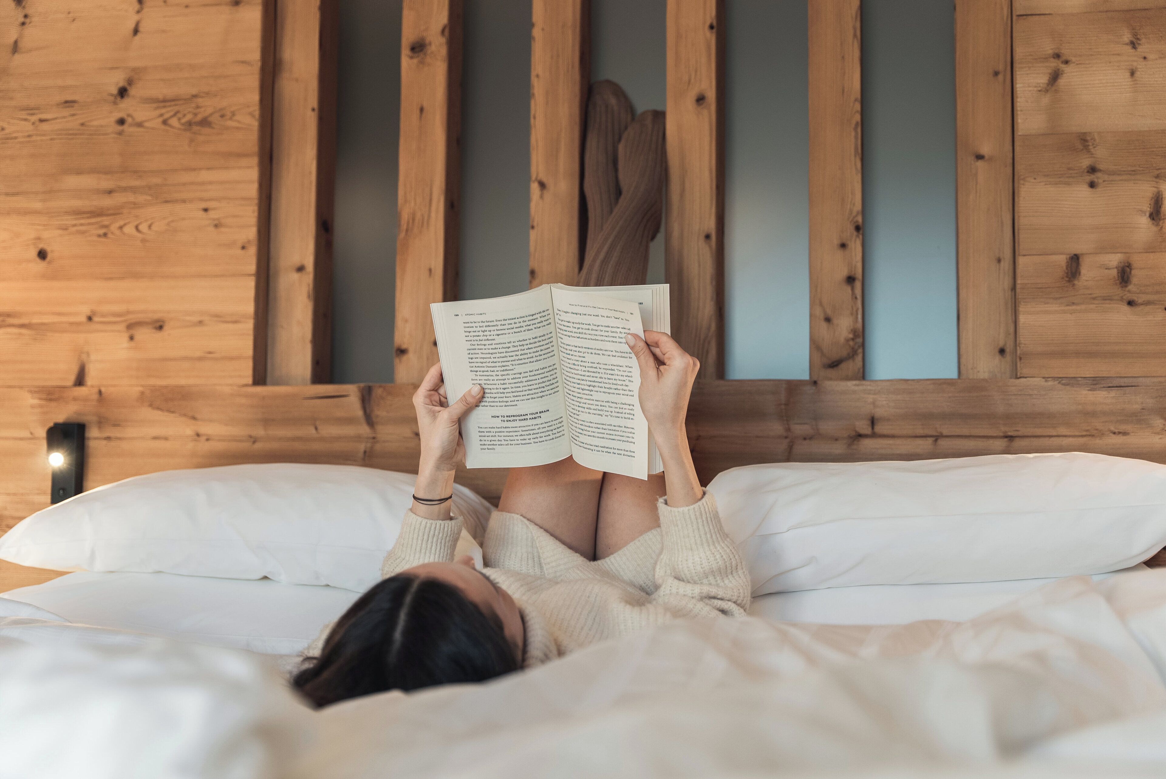 Una persona sdraiata su un letto con un libro, in una stanza dalle pareti rivestite in legno.