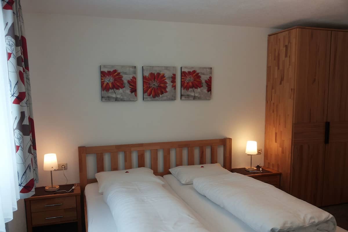A double bed with white bedding, two bedside lamps, a wooden headboard, and a wardrobe.