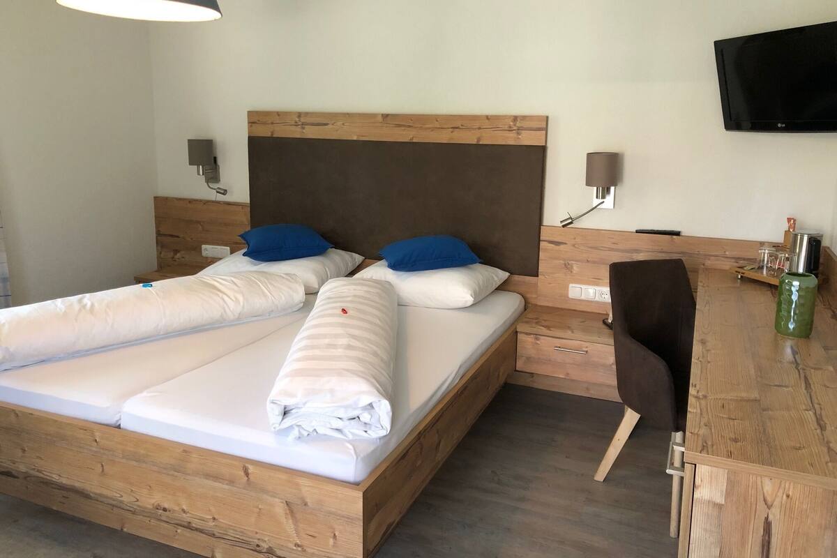 A double bed with a wooden headboard, two blue pillows, a wooden nightstand with a lamp, a TV mounted on the wall, and a wooden desk with a chair.
