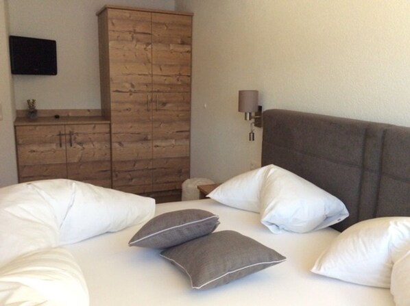 Un dormitorio con cama, almohadas, un armario de madera, un televisor de pantalla plana y una lámpara de noche.
