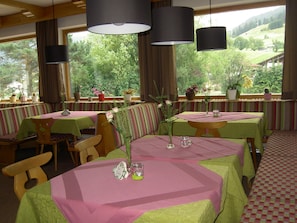 A restaurant with multiple tables and booths, each covered with green and pink tablecloths. There are potted plants and flowers on the windowsills, and the view includes green hills and trees. - Lusspark (Lermoos)