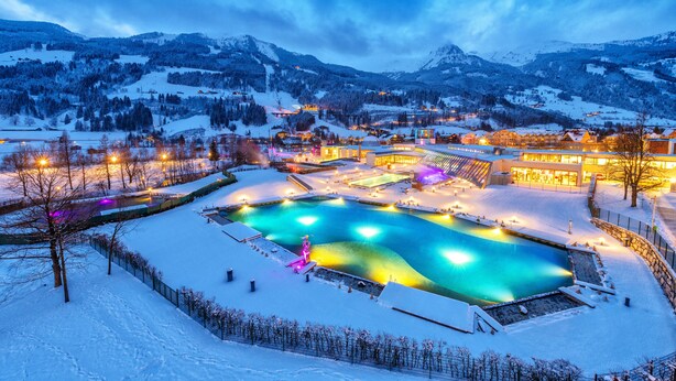 A snow-covered landscape with a large pool area illuminated by lights, surrounded by buildings and mountains in the background. - Hotel Germania Gastein (Bad Hofgastein)