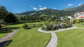 A grassy area with a gravel path, a small rock garden, and a view of mountains in the background. - Hotel Germania Gastein (Bad Hofgastein)