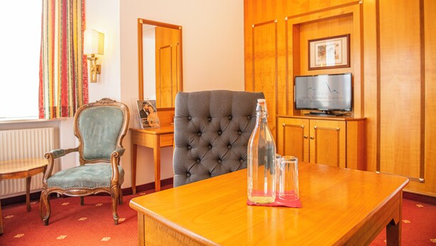 A hotel room with a wooden panel wall, a television, a tufted armchair, a wooden table, and a vintage armchair. - Hotel Germania Gastein (Bad Hofgastein)