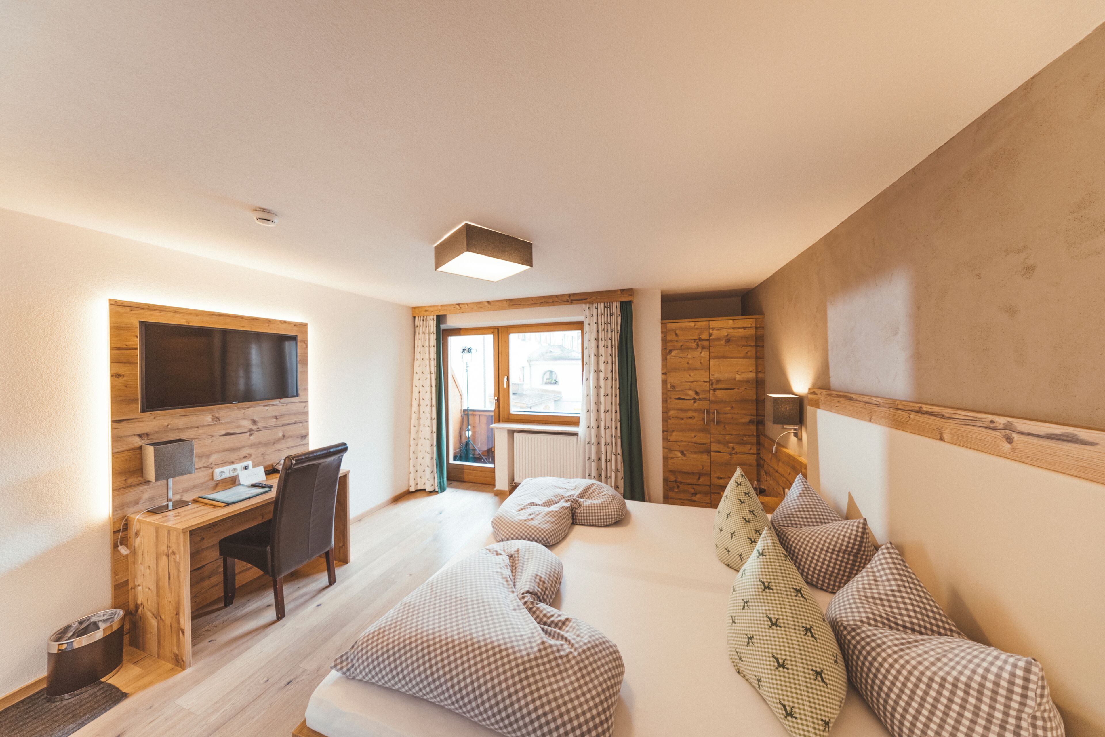 Ein Hotelzimmer mit Holzschreibtisch, einem Bett mit kariertem Bettbezug, einem an der Wand befestigten Fernseher und einem Fenster mit Vorhängen.