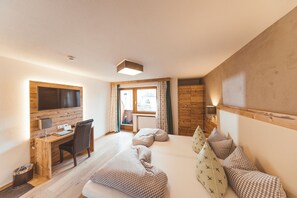 A hotel room with a wooden desk, a bed with checkered bedding, a television mounted on the wall, and a window with curtains. - Hubertushof (Lermoos)