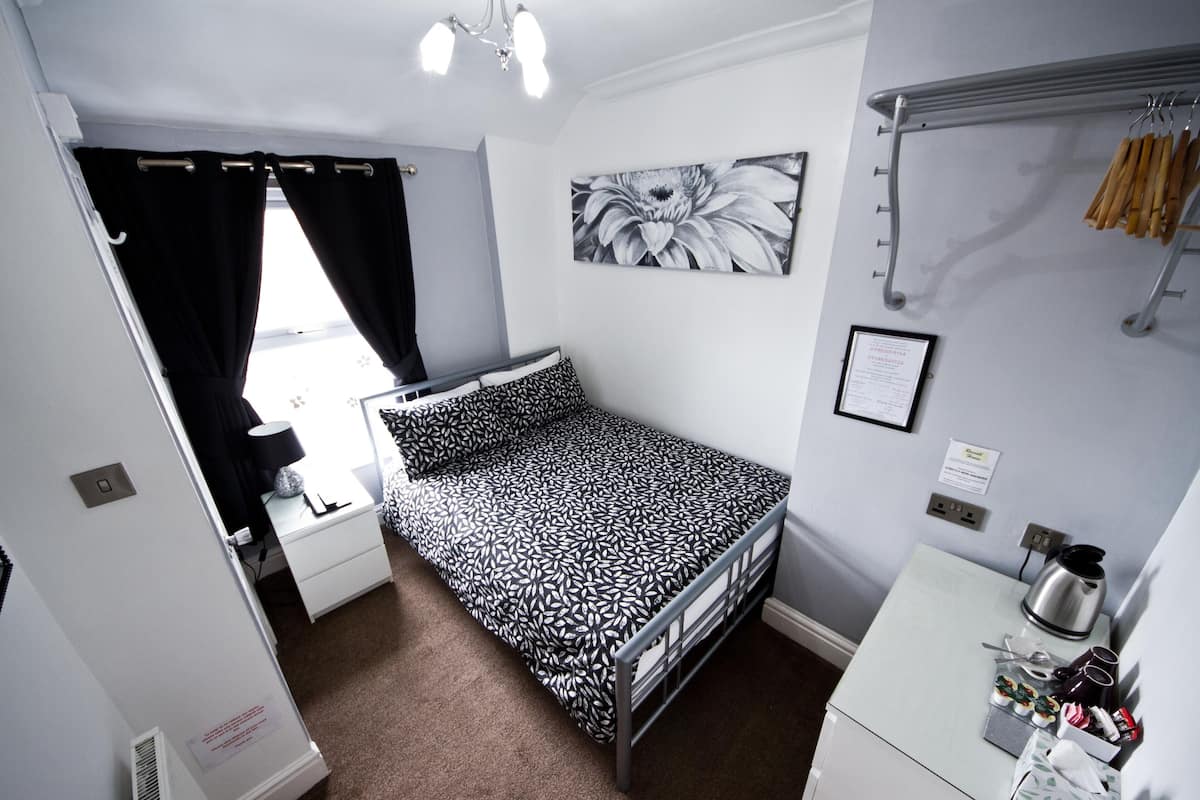 A compact, well-lit room with a single bed, a small desk with a kettle, and a wall-mounted drying rack.