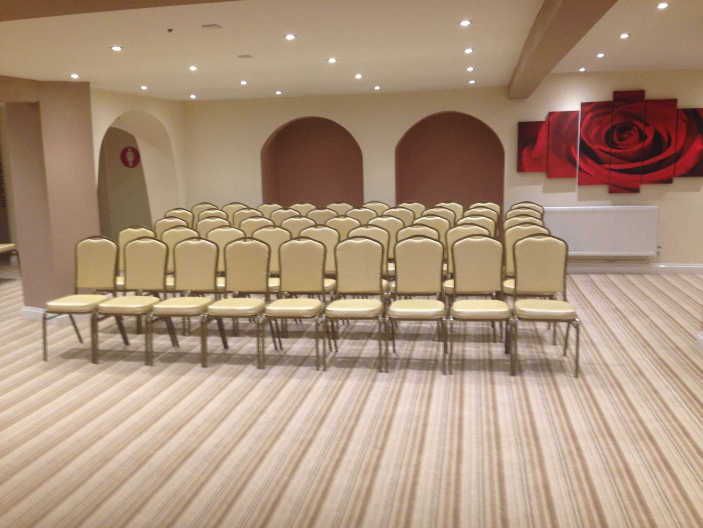 Sala de conferências com fileiras de cadeiras, piso coberto de carpete e parede com uma grande pintura de rosa vermelha.