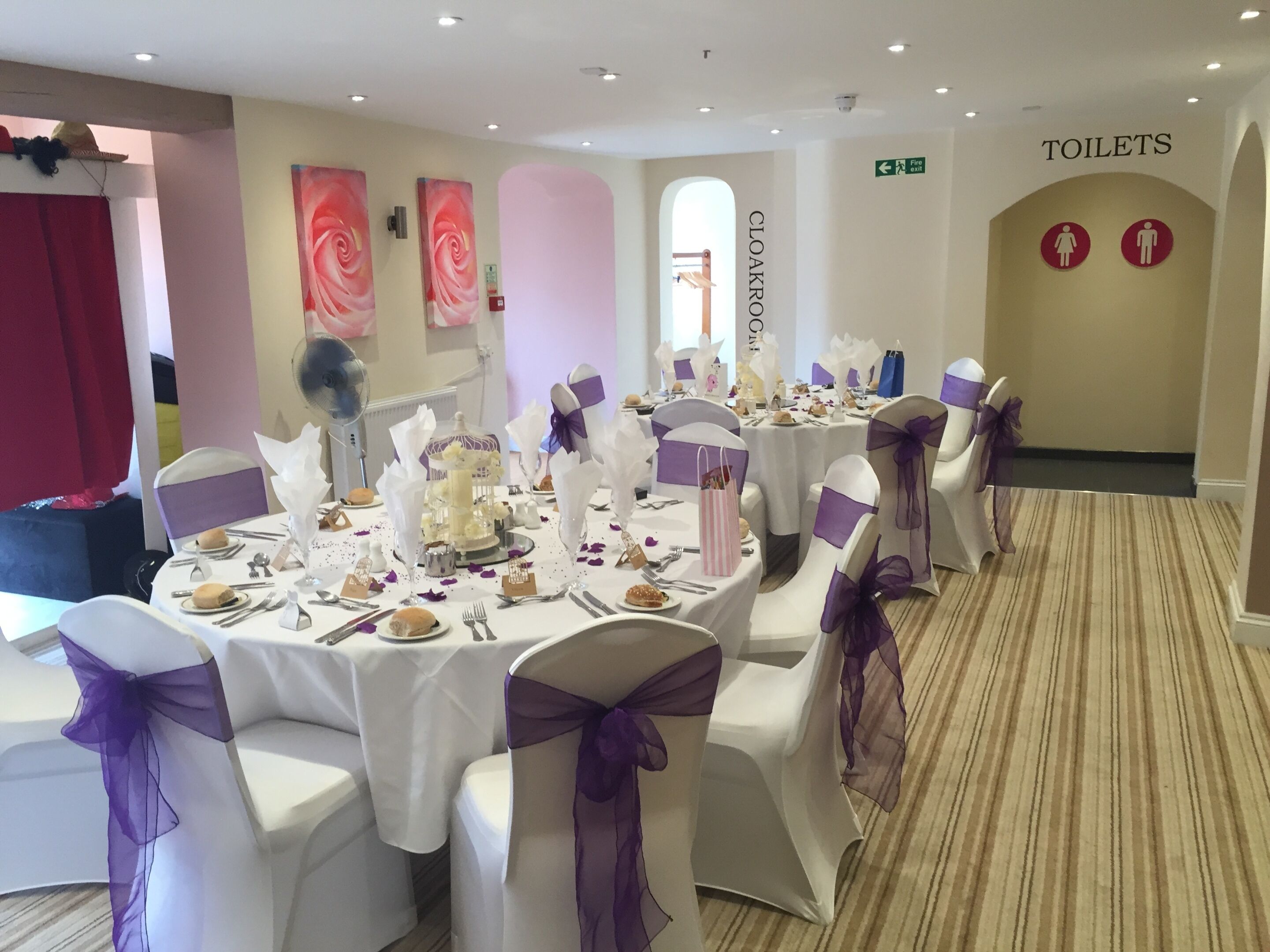 Un salón de banquetes con mesas redondas, manteles blancos y cintas moradas en las sillas.