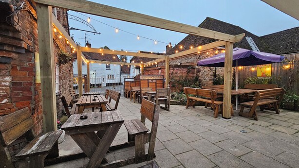 An outdoor patio with wooden tables and benches, string lights, and a brick wall. - The Crown Inn (Marlborough)