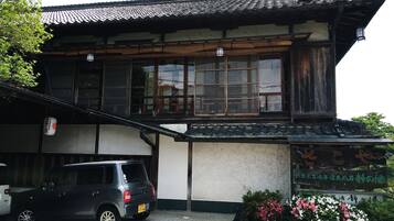 A traditional building with a tiled roof and wooden structure, a parked car, and a sign with Japanese characters.