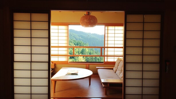 Una habitación tradicional japonesa con suelo de tatami, una mesa de madera baja, un banco acolchado y un amplio ventanal con vistas a una montaña boscosa.