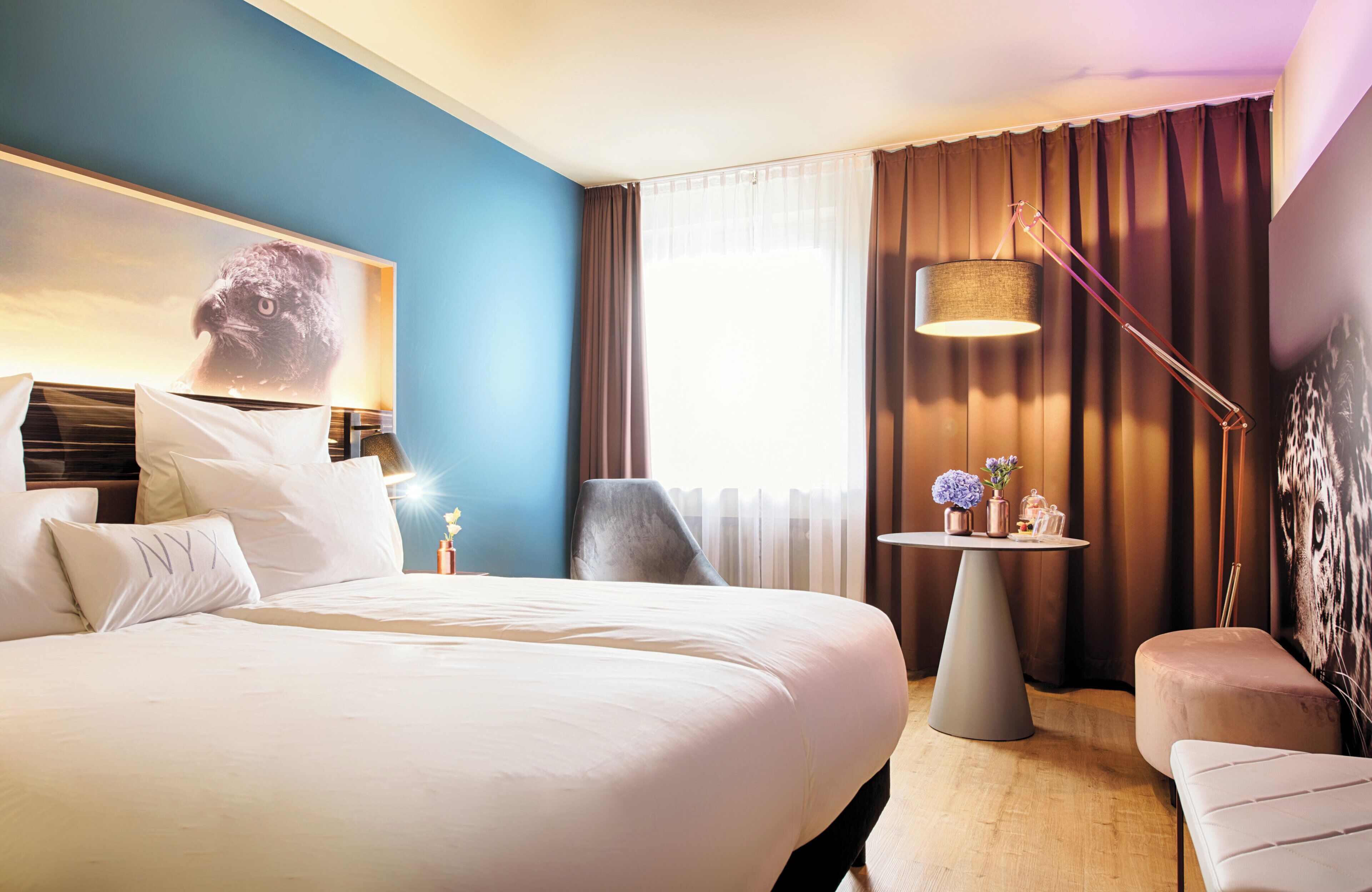 Habitación de hotel con una cama grande, una mesita de noche con lámpara, una silla y una pequeña mesa con flores.