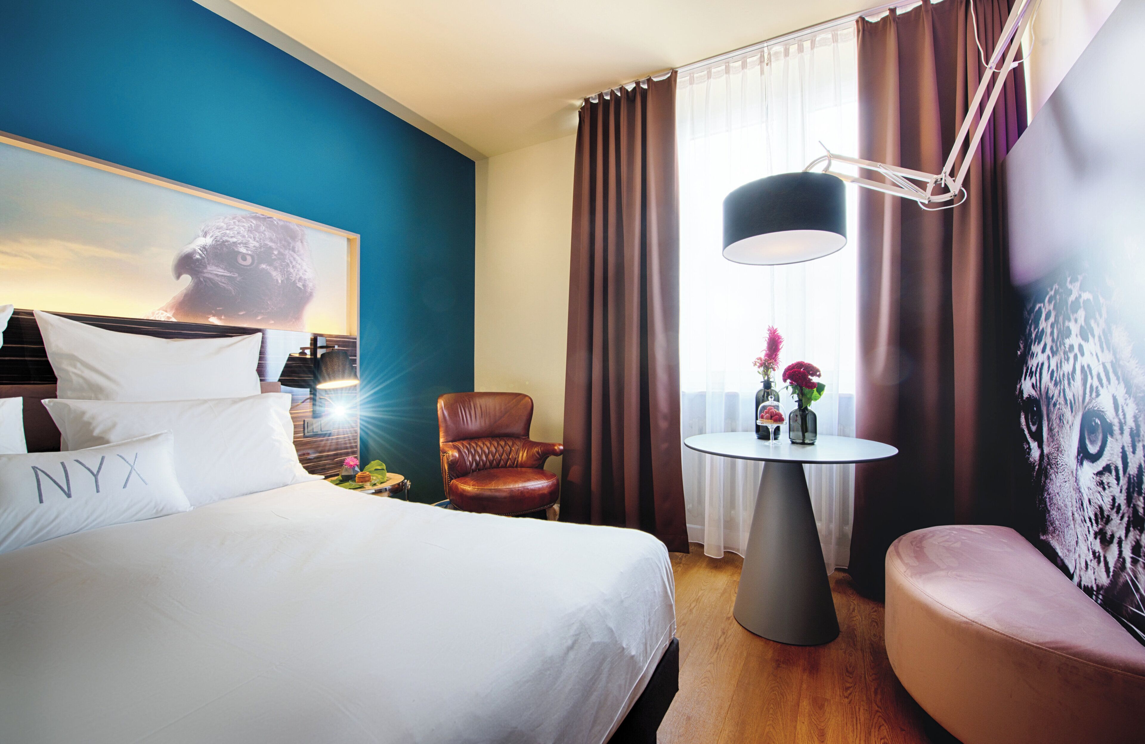 Ein Hotelzimmer mit einem großen Bett, einem Ledersessel, einem kleinen runden Tisch mit einer Vase und einem Tiergemälde an der Wand.