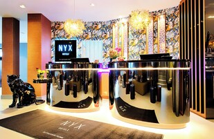 Balcão de recepção do NYX Hotels com design moderno, incluindo uma estátua de pantera negra, uma tela digital e um painel de parede com padrão floral.