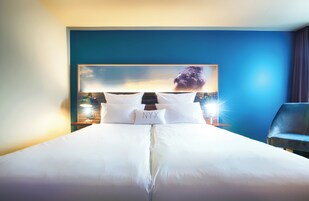 Una habitación de hotel con una cama grande, un cabecero con una pintura de un paisaje urbano, dos lámparas de noche y una silla.