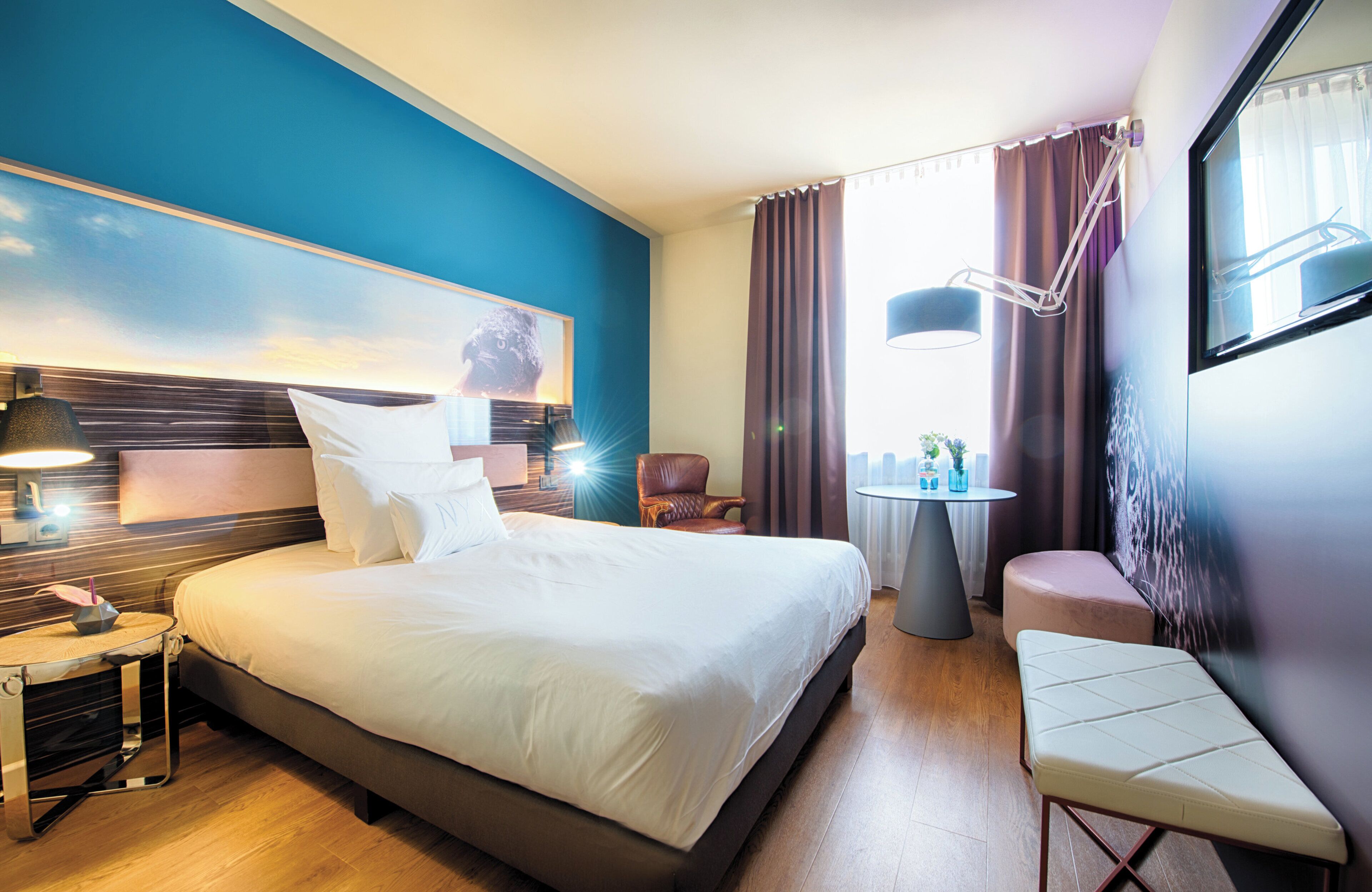Habitación de hotel con una cama grande, lámparas de noche, una mesita con lámpara, una silla y un banco.