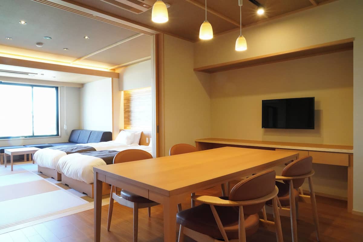 A modern hotel room with a wooden dining table, comfortable seating, a flat-screen TV, and a large window providing natural light.