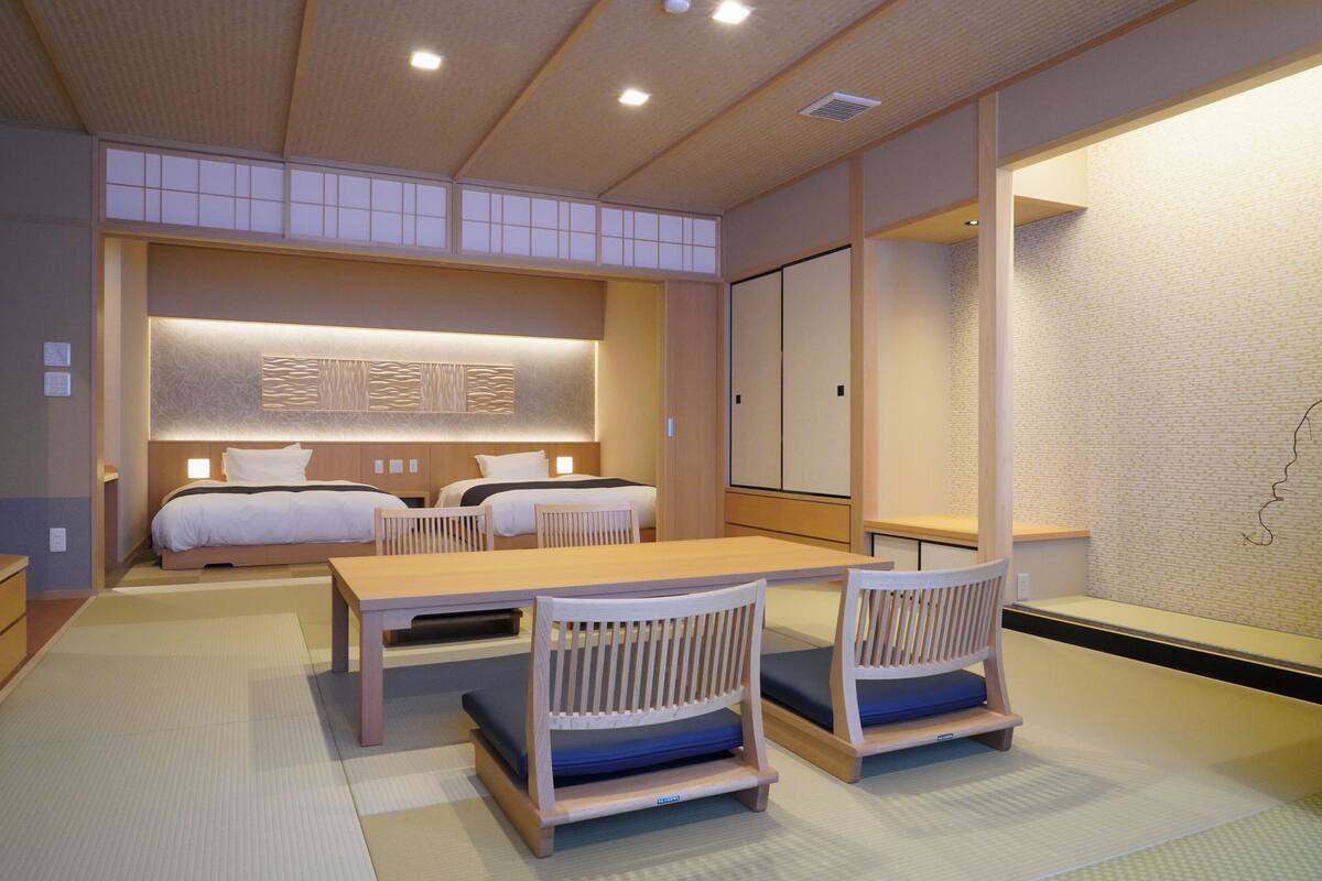 A hotel room with a traditional Japanese aesthetic, featuring tatami flooring, a low wooden table with chairs, a bed with a headboard, and sliding doors.