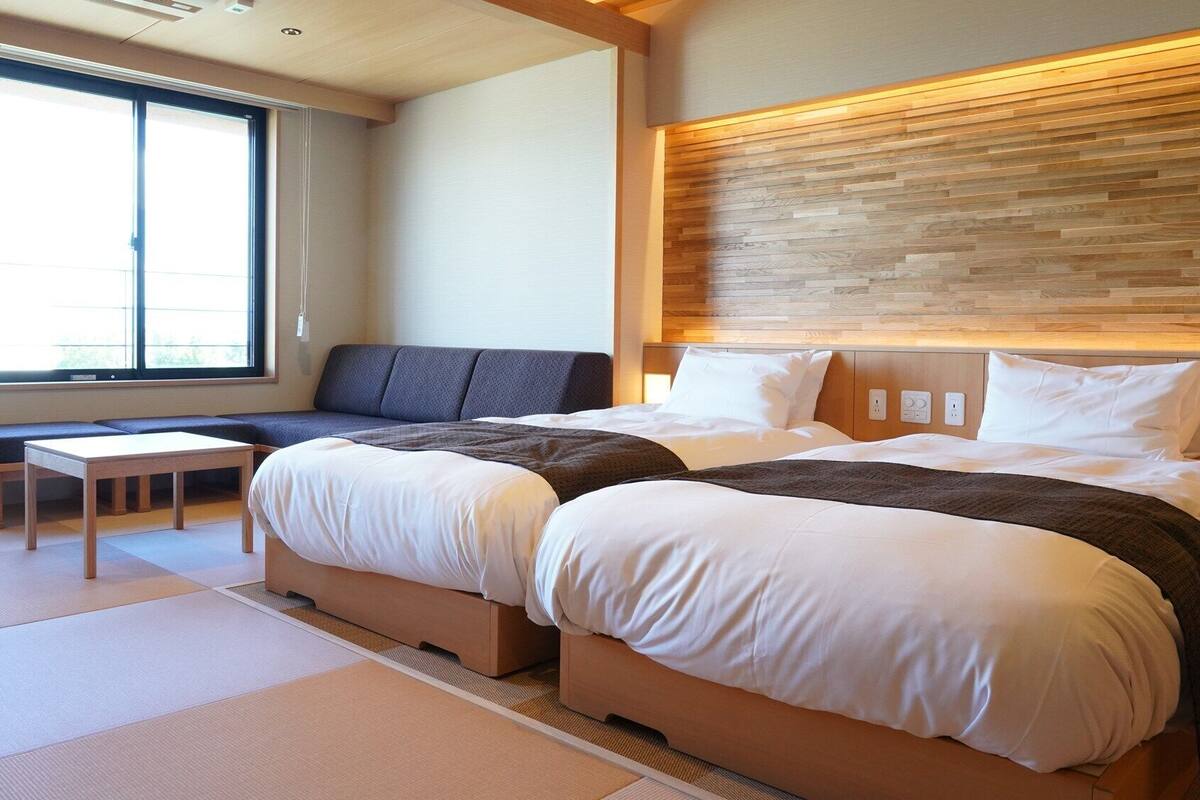 A hotel room with a large bed, a sofa, a small table, and a window with blinds.