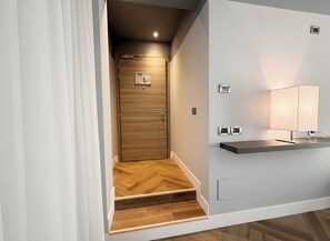 Ein modernes Hotelzimmer mit Holztür, einem kleinen Schreibtisch mit Lampe und einem Parkettboden mit Zickzackmuster.
