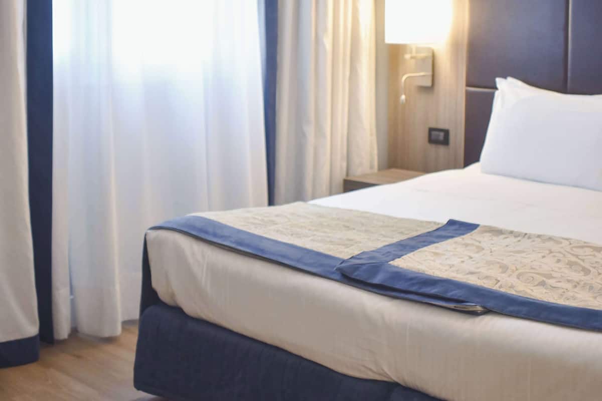 A hotel room with a large bed, a nightstand with a lamp, and a window with curtains.