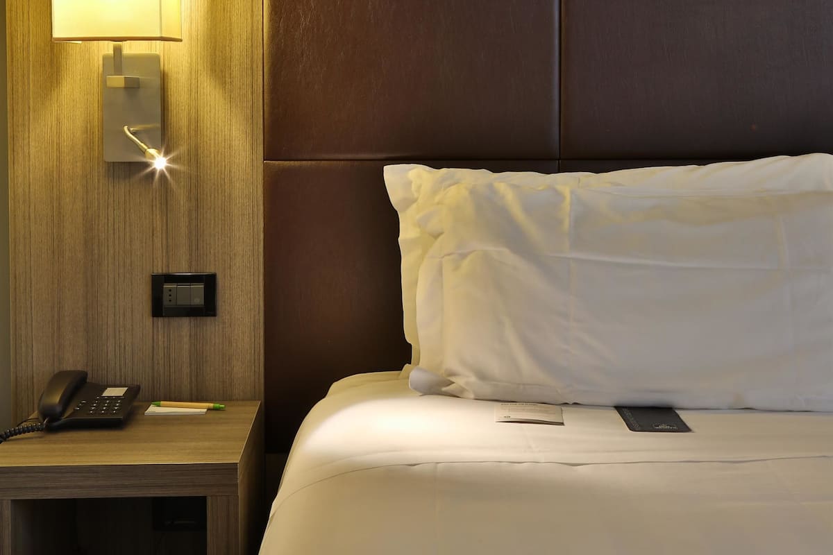 A hotel room with a bed, a bedside table with a phone, a lamp, and a wall-mounted control panel.