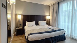 Amplia habitación de hotel con una cama grande, mesitas de noche, un espejo y vistas al exterior a través de cortinas translúcidas.