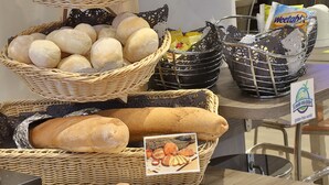 Una exhibición de panes y pasteles variados, incluyendo panes de molde y bollos, dispuestos sobre una barra con cestas y una tabla de cortar.