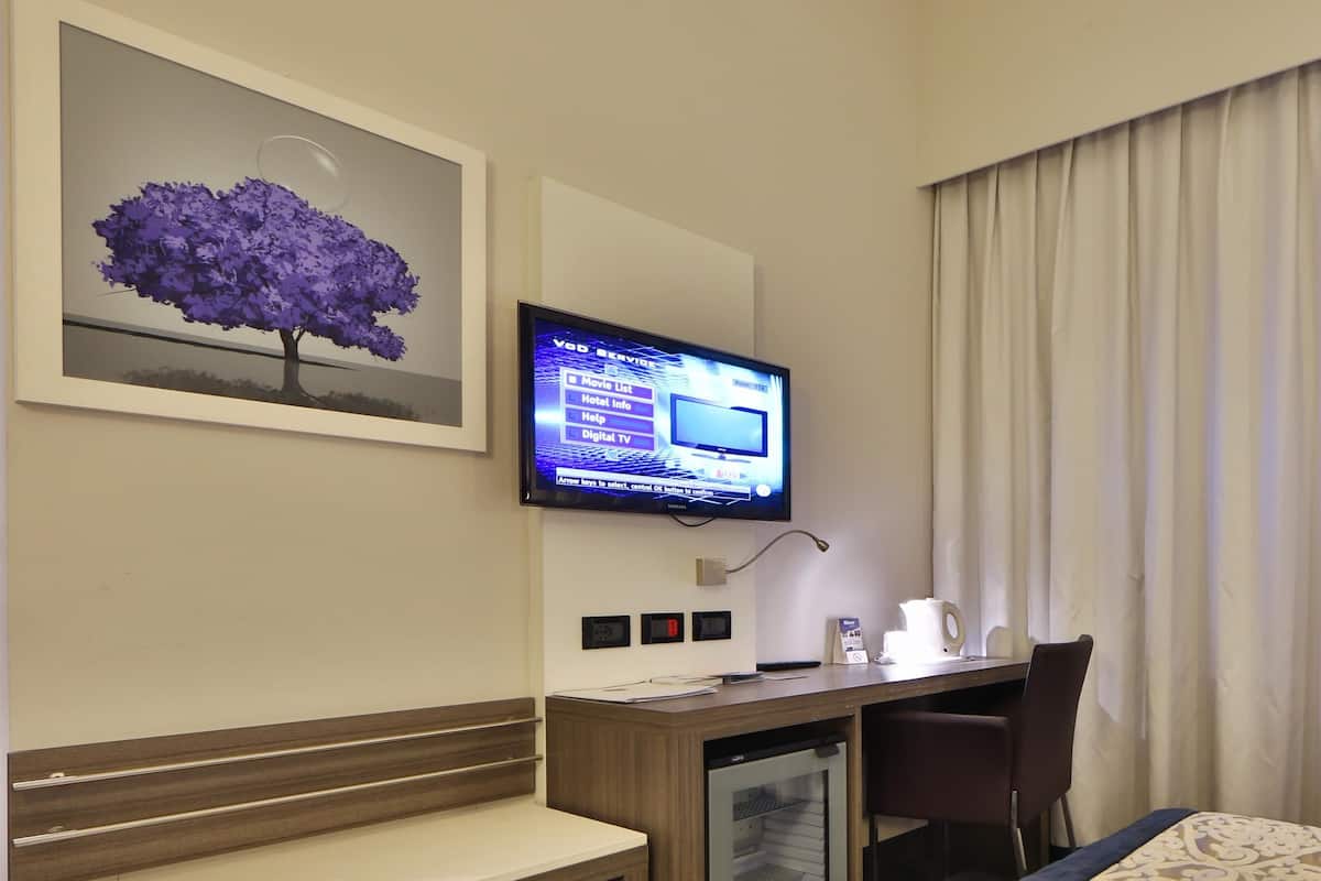 A hotel room with a desk, a television mounted on the wall, a small refrigerator, a bed with a patterned bedspread, and a framed picture of purple flowers.