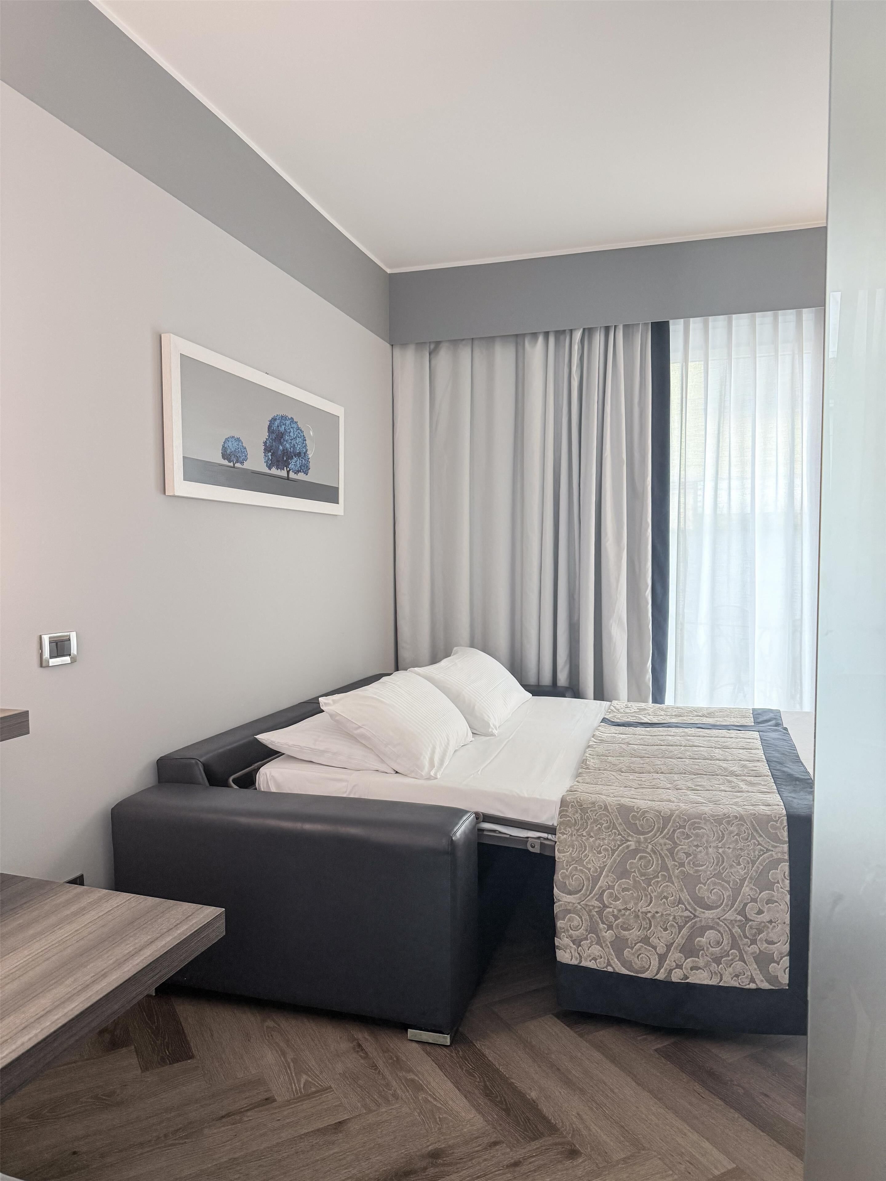 A modern hotel room with a bed, a bedside table, a chair, a window with curtains, and a framed picture on the wall.