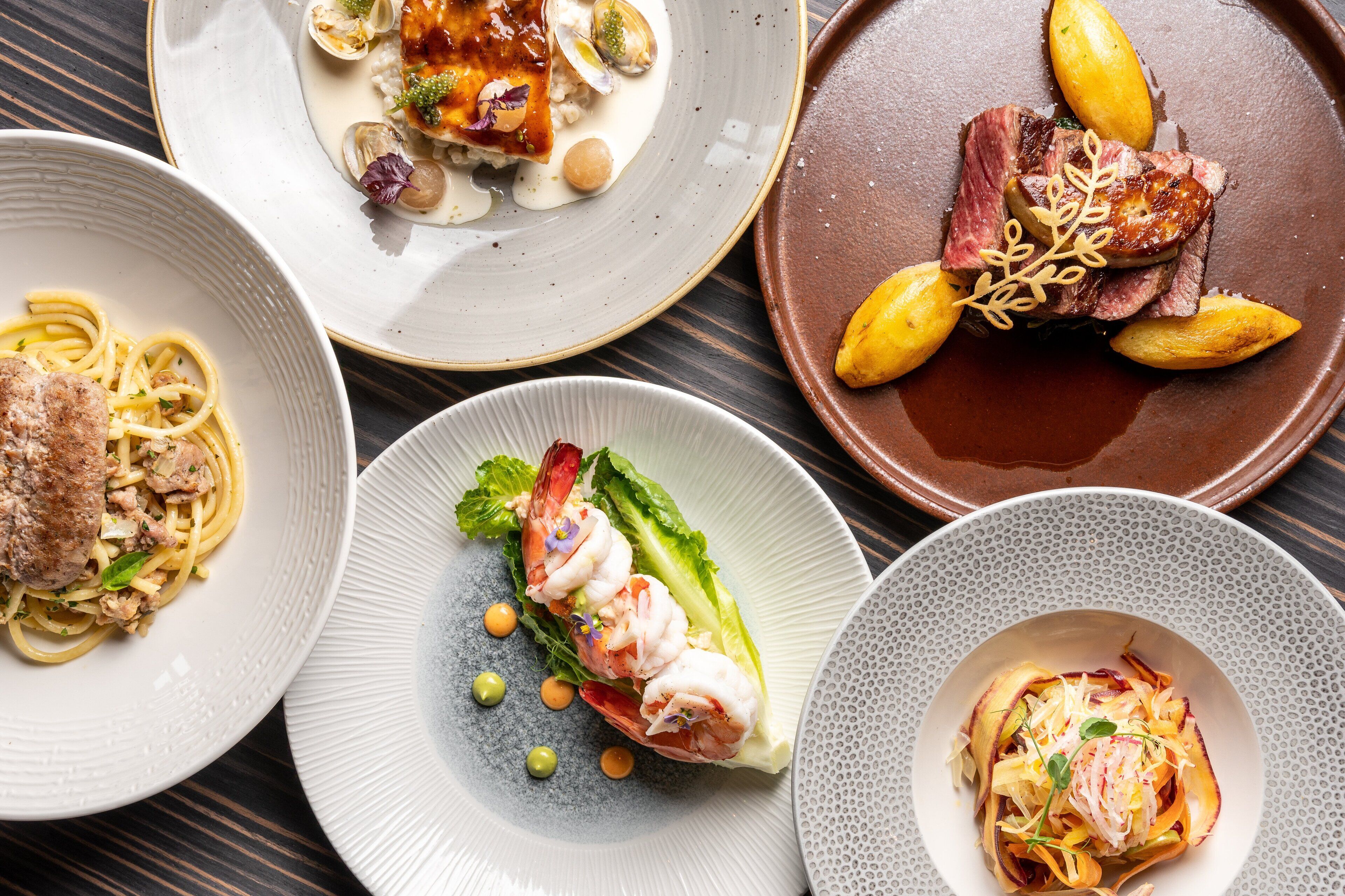 A variety of plated dishes including seafood, pasta, and steak, arranged on a wooden table.