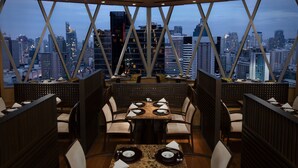 Un restaurant offrant une vue imprenable sur les toits d’une ville, avec un intérieur moderne et une grande table à manger centrale entourée de chaises.