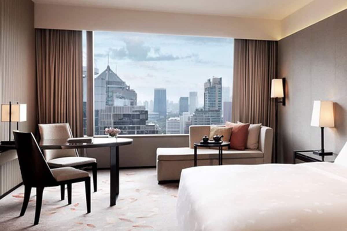 Habitación de hotel con una cama grande, un sofá, una silla, una mesita y vistas a la silueta del horizonte de la ciudad.