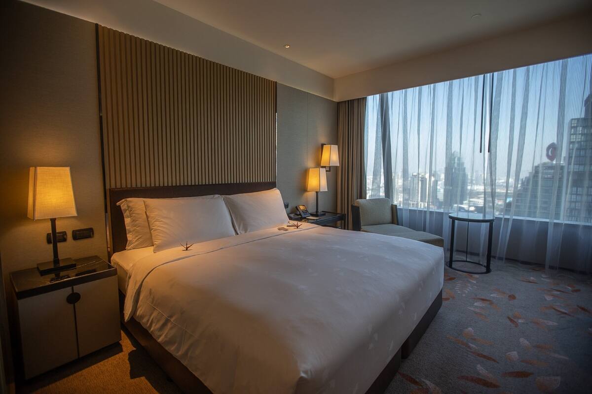 Habitación de hotel con una cama grande, dos lámparas de noche, una mesita de noche, una silla, una pequeña mesa y vistas a la ciudad a través de cortinas translúcidas.