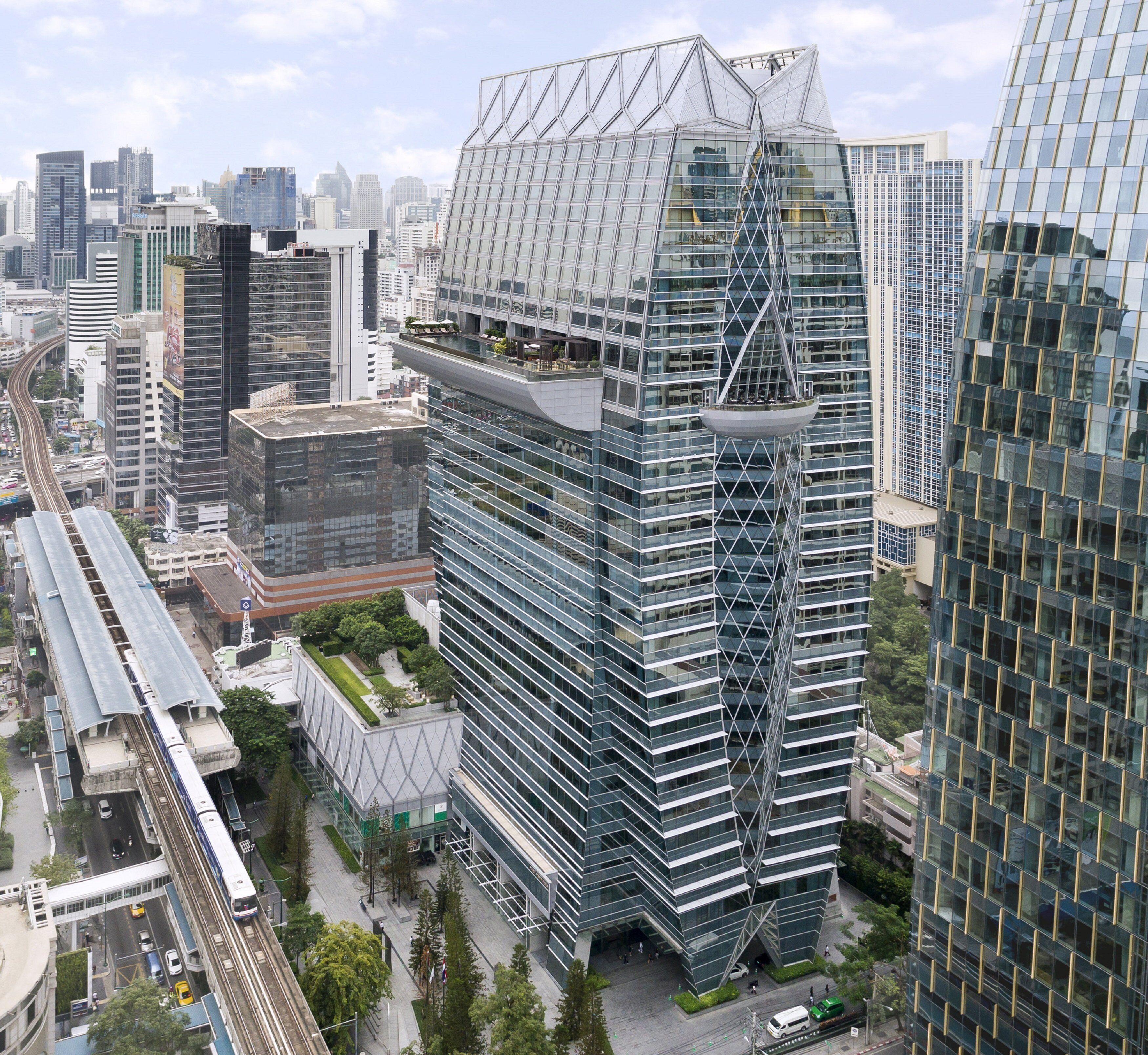 A modern high-rise building with a unique geometric glass facade, surrounded by other high-rise buildings and a train track.