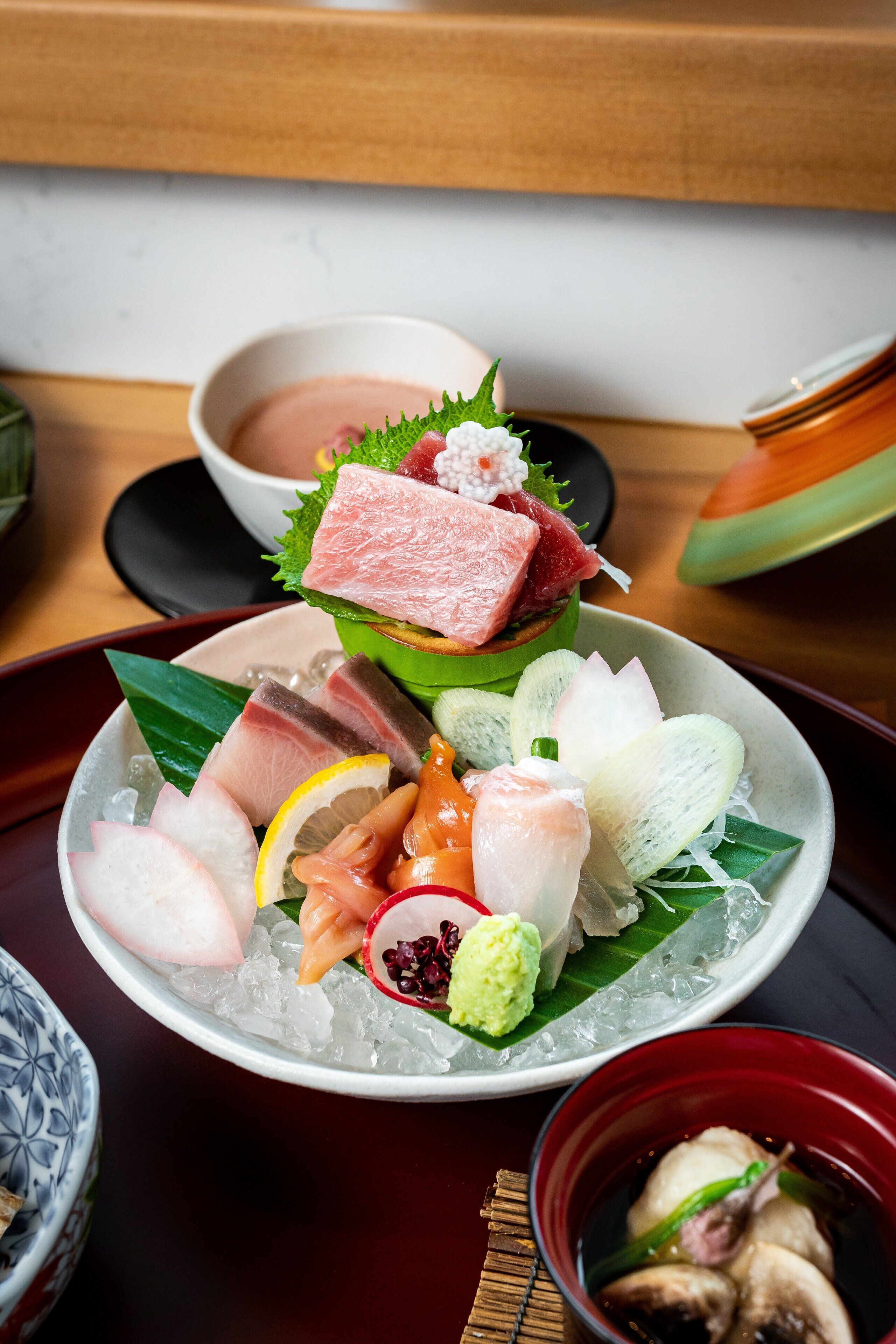 Comida tradicional japonesa con varios tipos de sashimi y sushi sobre una base de hielo, acompañada de un tazón pequeño de sopa y un plato decorativo.