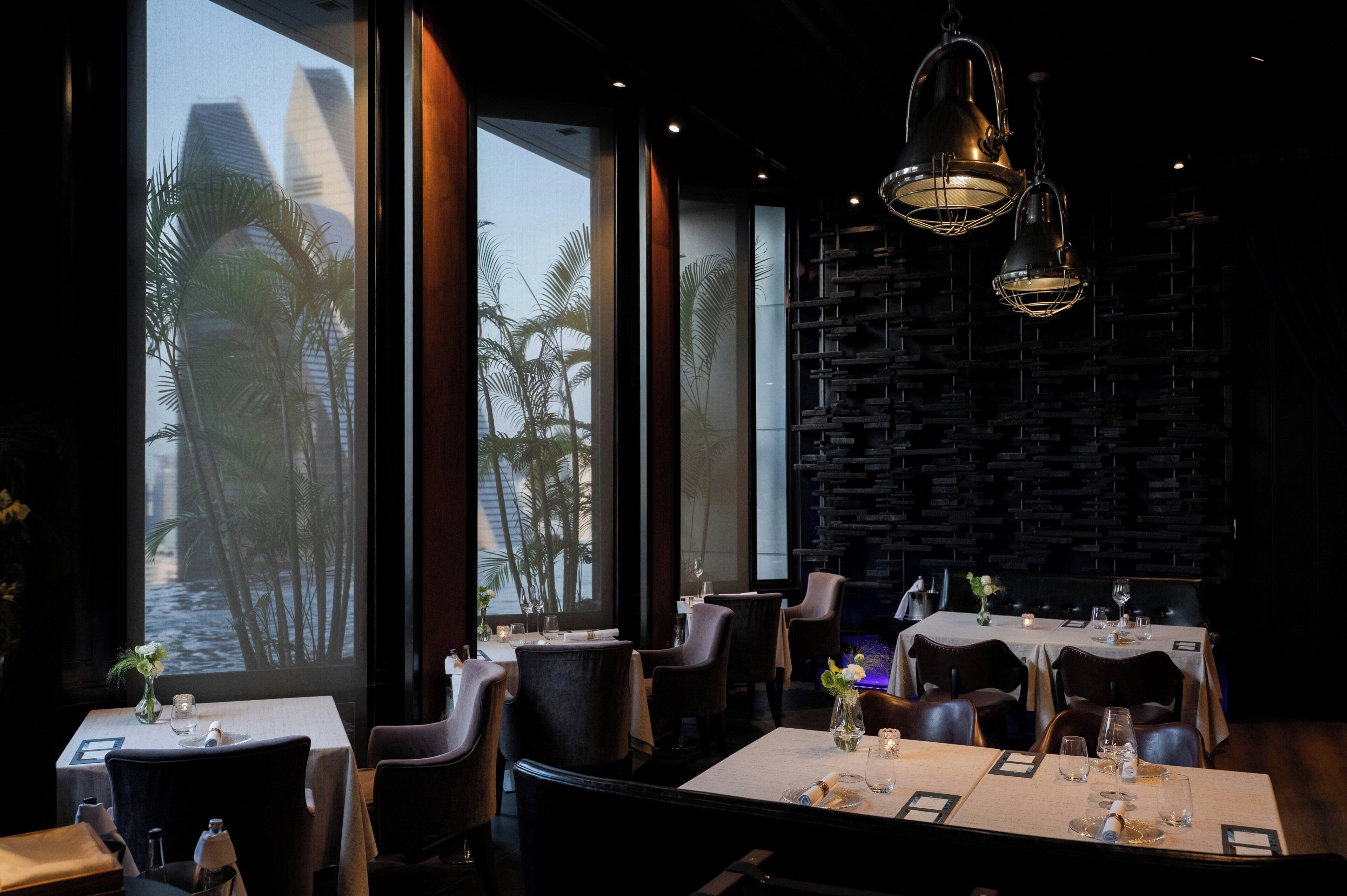 A restaurant interior with tables set for dining, large windows with a view of palm trees, and modern pendant lighting.