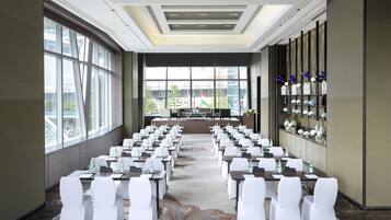 Une salle de conférence avec des rangées de tables et de chaises, de grandes fenêtres et un mur décoratif agrémenté d’étagères.