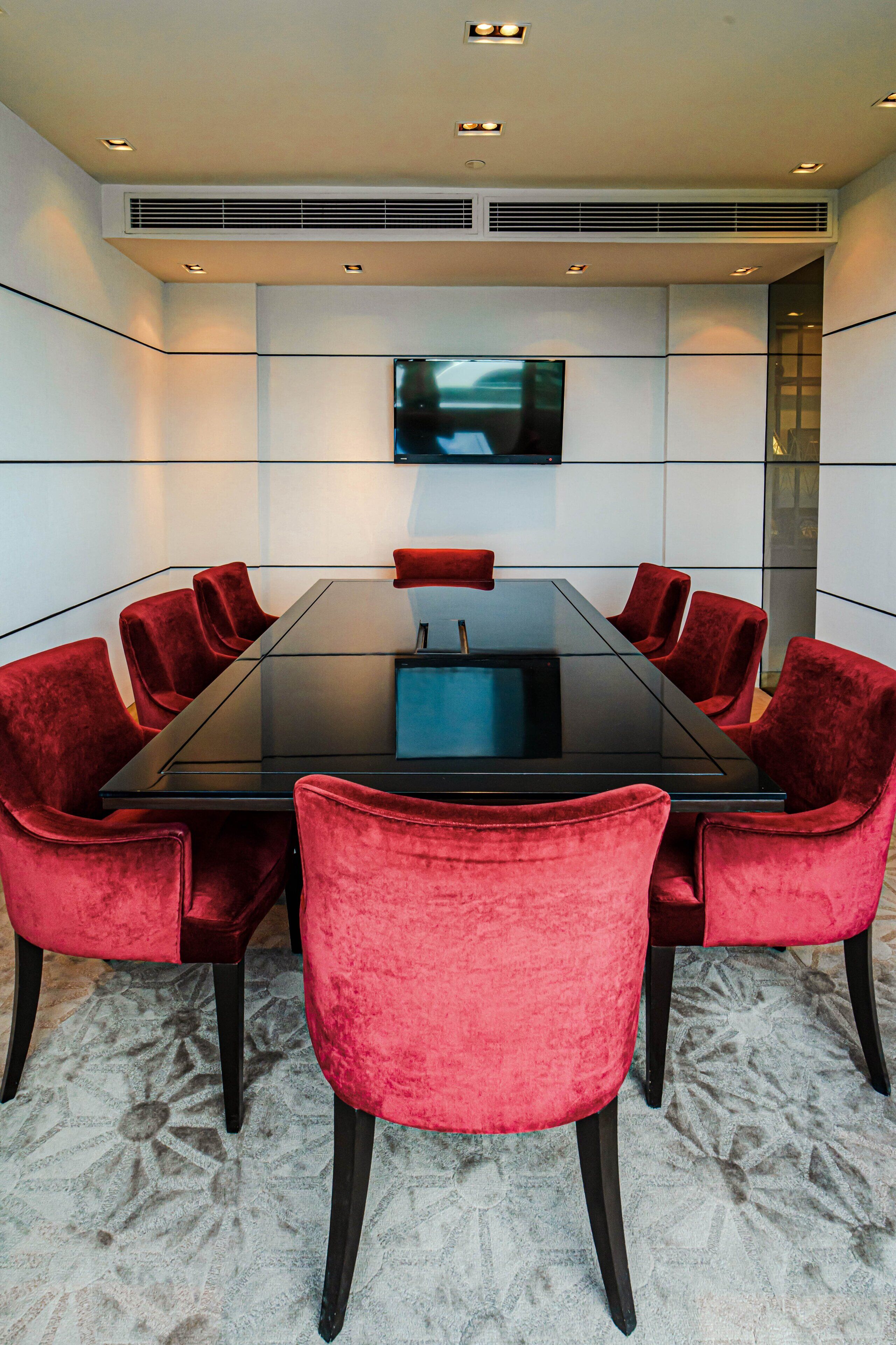 Een vergaderruimte met een glazen tafel, rode fluweelstoelen, een wandtelevisie en inbouwverlichting.