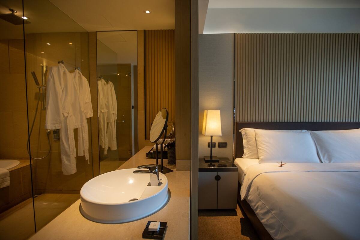 Habitación de hotel con ducha envuelta en vidrio, un lavabo redondo, una mesita de noche con lámpara y una cama con ropa blanca.