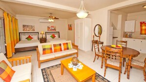 Ein hell erleuchtetes Wohnzimmer mit Sofa, Couchtisch, Esstisch und Stühlen. Ein Bett mit bunten Kissen, ein Deckenventilator und ein Blick in eine Küche durch einen offenen Durchgang sind ebenfalls zu sehen.