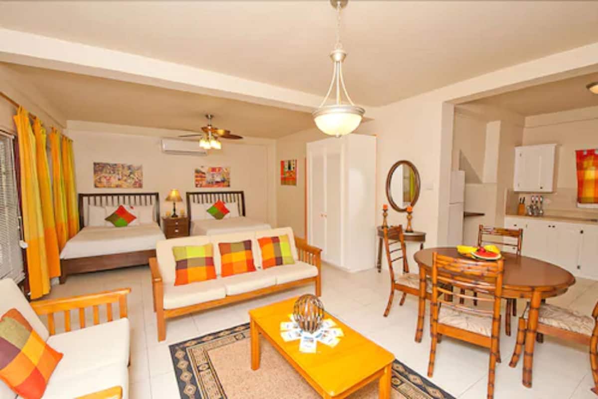 A well-lit living room with a sofa, coffee table, dining table, and chairs. There is a bed with colorful pillows, a ceiling fan, and a kitchen area visible through an open doorway.