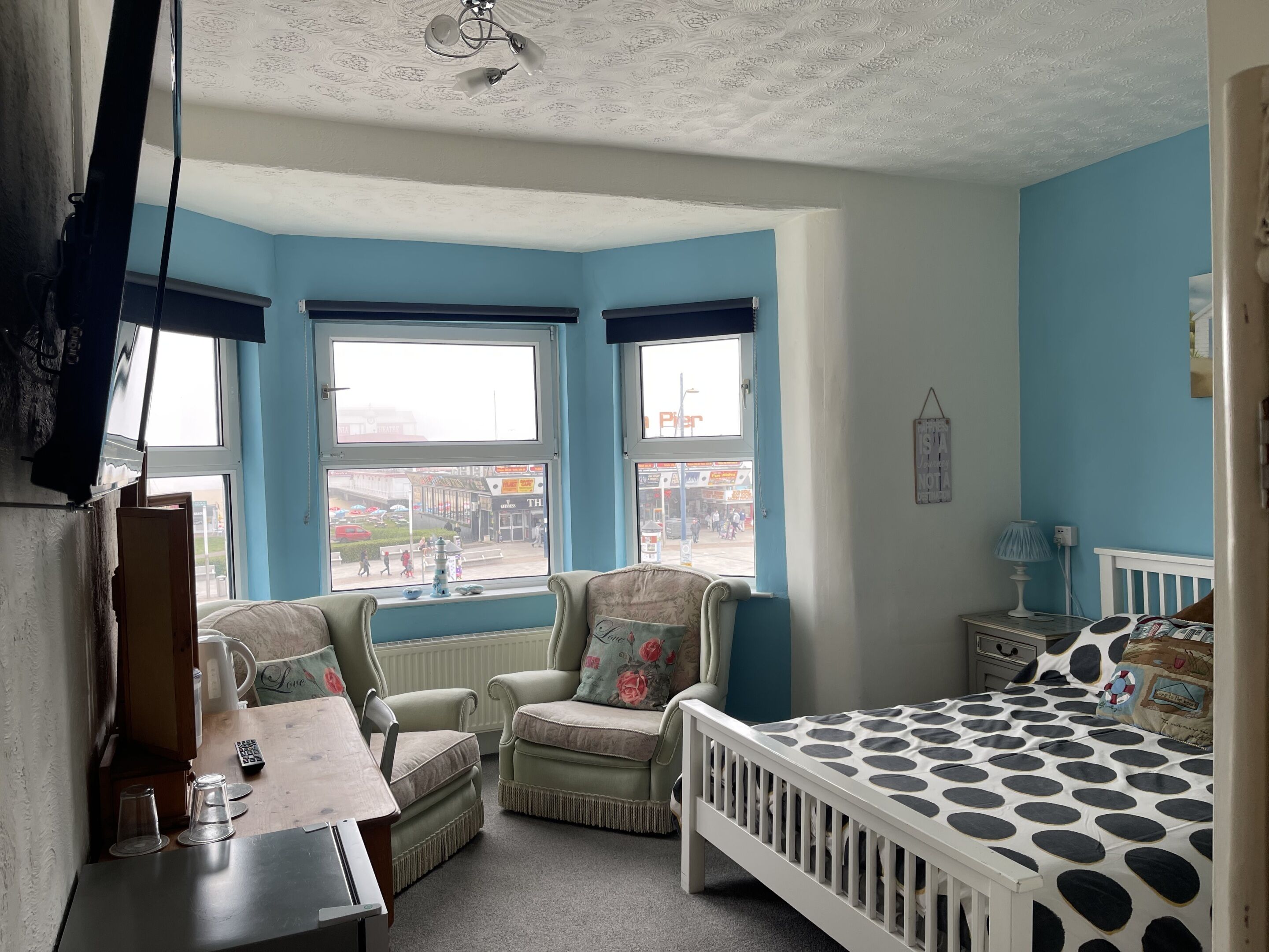 A bedroom with a bay window, two armchairs, a crib, a desk with a lamp, and a view of the street.